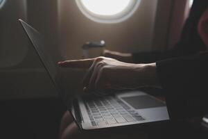Blonde female tourist checking incoming notification on smartphone sitting on seat of airplane with netbook.Young businesswoman share media from telephone on laptop computer during plane flight photo