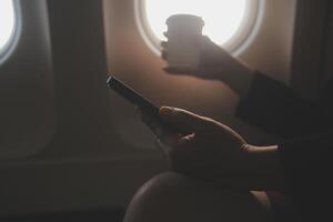 Blonde female tourist checking incoming notification on smartphone sitting on seat of airplane with netbook.Young businesswoman share media from telephone on laptop computer during plane flight photo