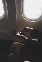 Blonde female tourist checking incoming notification on smartphone sitting on seat of airplane with netbook.Young businesswoman share media from telephone on laptop computer during plane flight photo