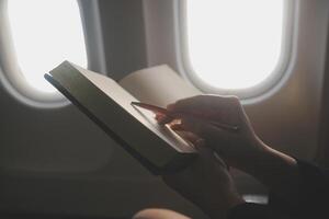 Blonde female tourist checking incoming notification on smartphone sitting on seat of airplane with netbook.Young businesswoman share media from telephone on laptop computer during plane flight photo