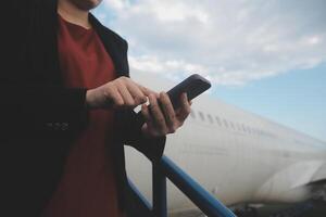 joven asiático mujer en internacional aeropuerto, utilizando móvil teléfono inteligente y comprobación vuelo a el vuelo información tablero foto