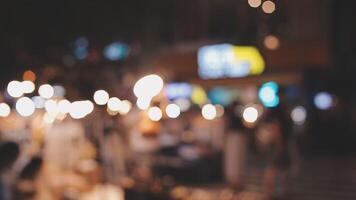 hermosa bokeh en un oscuro borroso antecedentes a noche en el caminando calle con el gente. el redondo vistoso bokeh brillar desde edificios y tiendas en el ciudad estilo de vida. resumen concepto. video