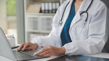 Serious female doctor using laptop and writing notes in medical journal sitting at desk. Young woman professional medic physician wearing white coat and stethoscope working on computer at workplace. video