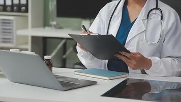 sérieux femelle médecin en utilisant portable et l'écriture Remarques dans médical journal séance à bureau. Jeune femme professionnel médical médecin portant blanc manteau et stéthoscope travail sur ordinateur à lieu de travail. video