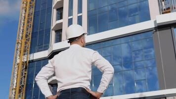 een architect ingenieur in een wit overhemd en helm staat met zijn terug naar de camera tegen de backdrop van een modern glas gebouw video