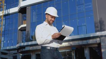 Engineer architect in a white shirt and helmet on the background of a modern glass building works making notes in a tablet video