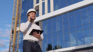 An engineer-architect in a white shirt and helmet on a background of a modern glass building holds a tablet and talks on a cell phone video