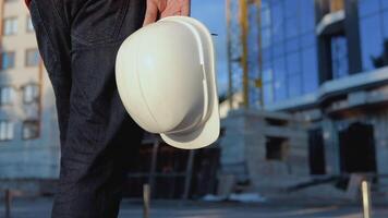 An engineer-architect in a white shirt and orange work vest stands against the backdrop of a modern glass building. Close-up view of a helmet in the architect's hand video