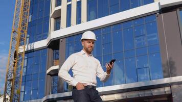 Ingenieur Architekt im ein Weiß Hemd und Helm auf das Hintergrund von ein modern Glas Gebäude spricht auf ein Zelle Telefon. modern Konstruktion video