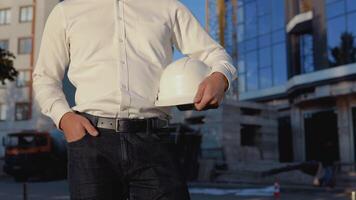 ingeniero en un blanco camisa en el antecedentes de un moderno vaso edificio y sostiene un un casco. de cerca ver de un casco en un del hombre mano video