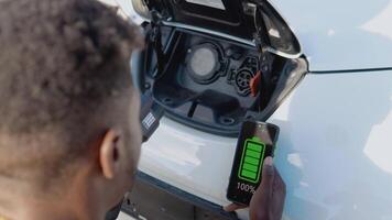 A dark-skinned male driver connects an electric car to the power system to charge the car battery and controls the charging process via his smartphone video