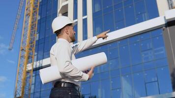 un ingénieur dans une blanc chemise et casque travaux sur le construction de une moderne verre bâtiment et détient une rouleau avec une dessin de le projet video