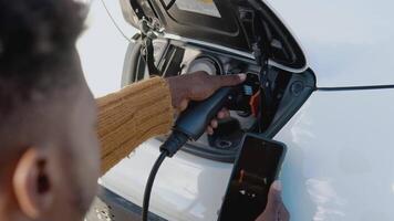 African american male driver connects an electric car to the power system to charge the car battery and controls the charging process via his smartphone video