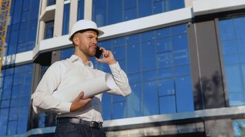 The engineer-architect holds a roll with a drawing of the project and talks on a cell phone. An engineer in a white shirt and helmet works on the construction of a modern glass building video