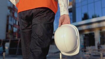 An engineer-architect in a white shirt and orange work vest walks in the direction of a modern glass building. Close-up view of a helmet in the architect's hand video