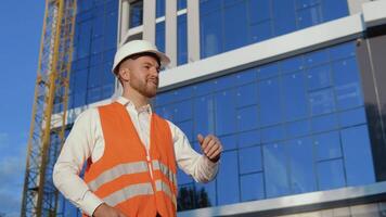 a engenheiro-arquiteto dentro uma branco camisa, capacete e laranja trabalhos colete carrinhos contra a pano de fundo do uma moderno vidro construção video