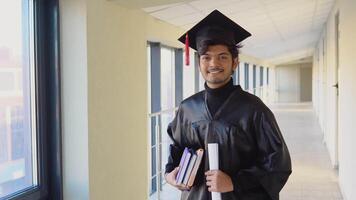 Indien diplômé dans manteau des stands avec une diplôme et livres dans sa mains et sourit. un important événement. Jeune spécialiste video