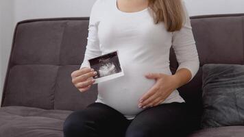 A pregnant fair-skinned woman strokes her tummy and shows a photo of the ultrasound diagnosis of the fetus video