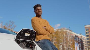 A dark-skinned driver is standing near a white electric car while charging the battery video