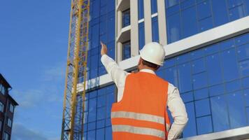 un ingeniero-arquitecto en un blanco camisa, casco y naranja trabajo chaleco soportes con su espalda a el cámara en contra el fondo de un moderno vaso edificio y supervisa el construcción proceso video