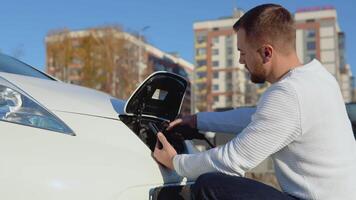 A fair-skinned male driver connects an electric car to the power system to charge the car battery and controls the charging process via his smartphone video