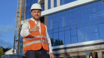 un ingeniero-arquitecto en un blanco camisa, casco y naranja trabajo chaleco soportes en contra el fondo de un moderno vaso edificio video