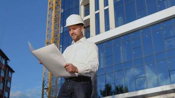 An engineer in a white shirt and helmet works on the construction of a modern glass building. Architect engineer reads project drawings video