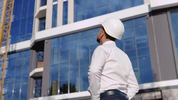 un architecte ingénieur dans une blanc chemise et casque des stands avec le sien retour à le caméra contre le toile de fond de une moderne verre bâtiment video