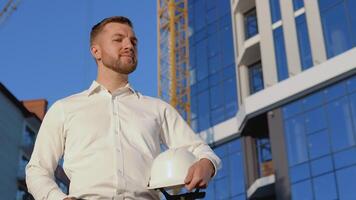 Architect engineer in a white shirt on the background of a modern glass building and holding a helmet video