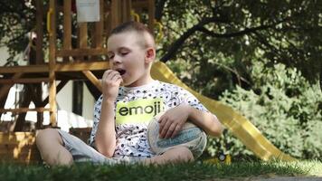 een tiener in korte broek, een t-shirt en blootsvoets zit Aan de gras in de park met een voetbal bal onder zijn oksel en een groen appel in zijn hand- video