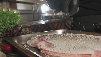 Chef in gloves salting and peppering fish lying on kitchen table surrounded by greens, tomatoes and vegetables video