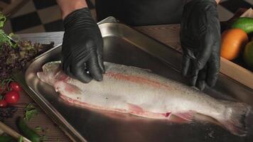 Chef in gloves salting fish on kitchen table among herbs, tomatoes and vegetables video