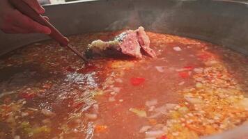 Chef mixes meat soup with vegetables in a large cauldron with a slotted spoon video