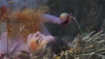 Young woman in autumn designer outfit of flowers, leaves and spikelets lying in the grass among scattered apples video