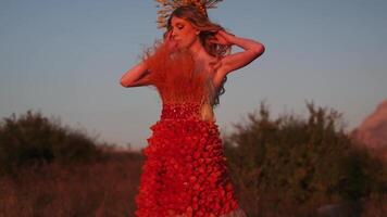 A young woman in an autumn outfit of flowers and grass and a headdress made of ears of corn stands against the background of the sunset in the mountains video