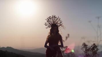 ung kvinna modell i designer huvudbonad tillverkad av spikelets och klänning tillverkad av blommor och gräs på de bakgrund av solnedgång i de berg. video