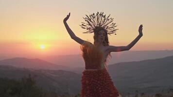 Jeune femelle modèle dans designer coiffure fabriqué de épillets et robe fabriqué de fleurs et herbe sur le Contexte de le coucher du soleil dans le montagnes. video