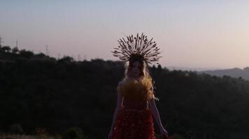 Film crew with a young female model in an autumn designer dress made of flowers and a headdress made of spikelets on the background of the sunset in the mountains. video