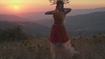 Jeune femelle modèle dans designer coiffure fabriqué de épillets et robe fabriqué de fleurs et herbe sur le Contexte de le coucher du soleil dans le montagnes. video