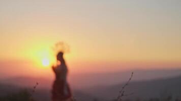 giovane femmina modello nel progettista copricapo fatto di spighette e vestito fatto di fiori e erba su il sfondo di tramonto nel il montagne. video