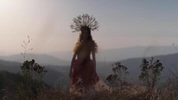 giovane femmina modello nel progettista copricapo fatto di spighette e vestito fatto di fiori e erba su il sfondo di tramonto nel il montagne. video