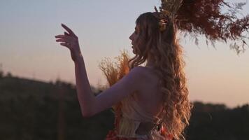 A young woman in a headdress made of spikelets and in an autumn dress of flowers holds dry branches with leaves in her hand on the background of sunset in the mountains video