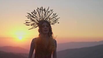 jong vrouw model- in ontwerper hoofdtooi gemaakt van aartjes en jurk gemaakt van bloemen en gras Aan de achtergrond van zonsondergang in de bergen. video