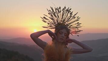 joven hembra modelo en diseñador tocado hecho de espiguillas y vestir hecho de flores y césped en el antecedentes de puesta de sol en el montañas. video
