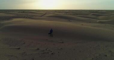 de Drönare flugor runt om en man i arab kläder gående genom de sand sanddyner av de öken- mot de bakgrund av de solnedgång. video