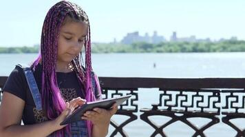 joven niña con rosado coletas usos un tableta en pie por el río. lento movimiento video