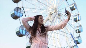 Young beautiful girl with long hair in a long dress makes selfie using a smartphone standing near the Ferris wheel. slow motion. video