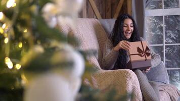 família Natal, Diversão festa, fique às lar, Novo ano celebração. uma mulher abre uma presente caixa enquanto sentado dentro uma cadeira às casa em Natal véspera video