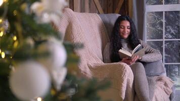 familie kerstmis, pret partij, blijven Bij huis, nieuw jaar viering. vrouw in gebreid trui leest een boek terwijl zittend in een stoel in de buurt de Kerstmis boom video