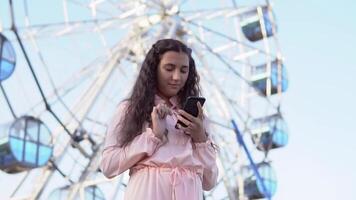 un' bellissimo ragazza usi un' Telefono mentre in piedi vicino il Ferris ruota. lento movimento video
